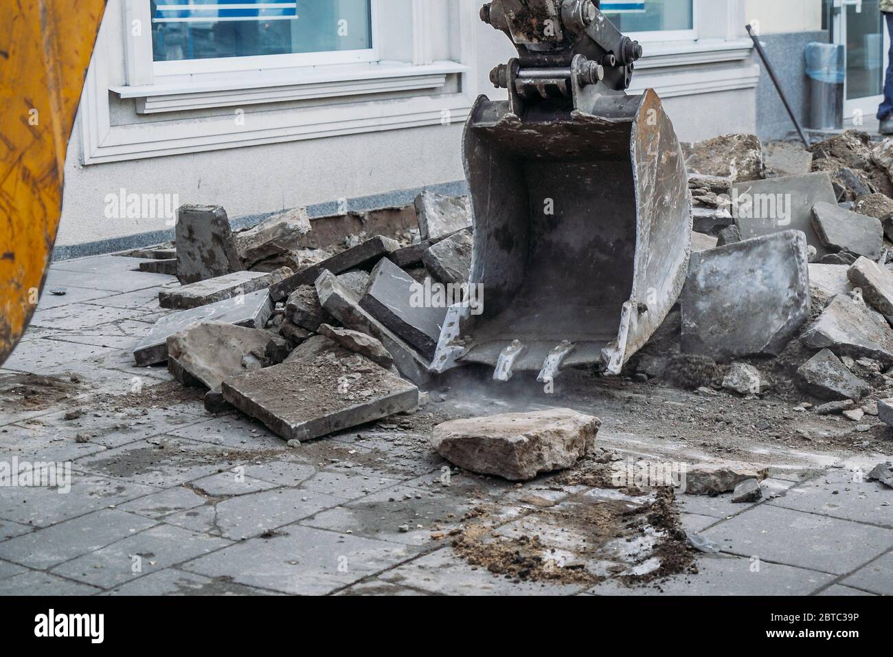 Excavator bucket breaks paving slabs. Sidewalk dismantling and repair work in city. Stock Photo