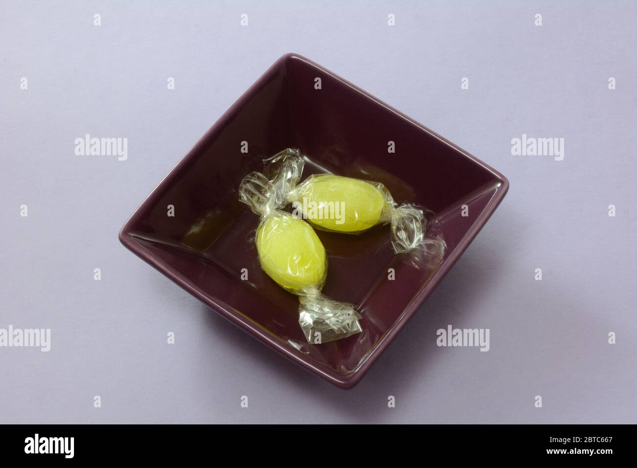 Sherbet lemon drop hard candy in plastic wrapper in purple square bowl on lavender background Stock Photo