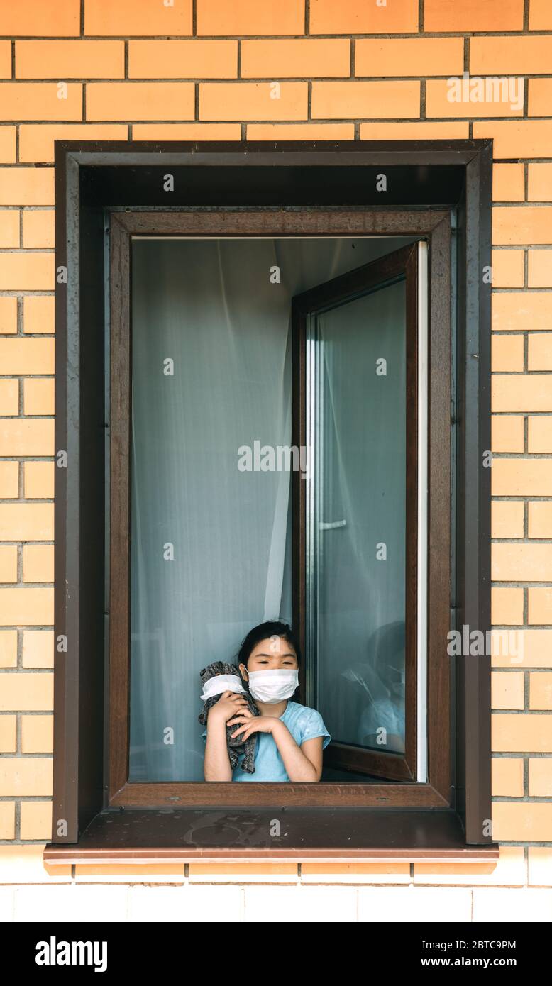 A girl in a mask with a stuffed toy stands at the open window of her house Stock Photo