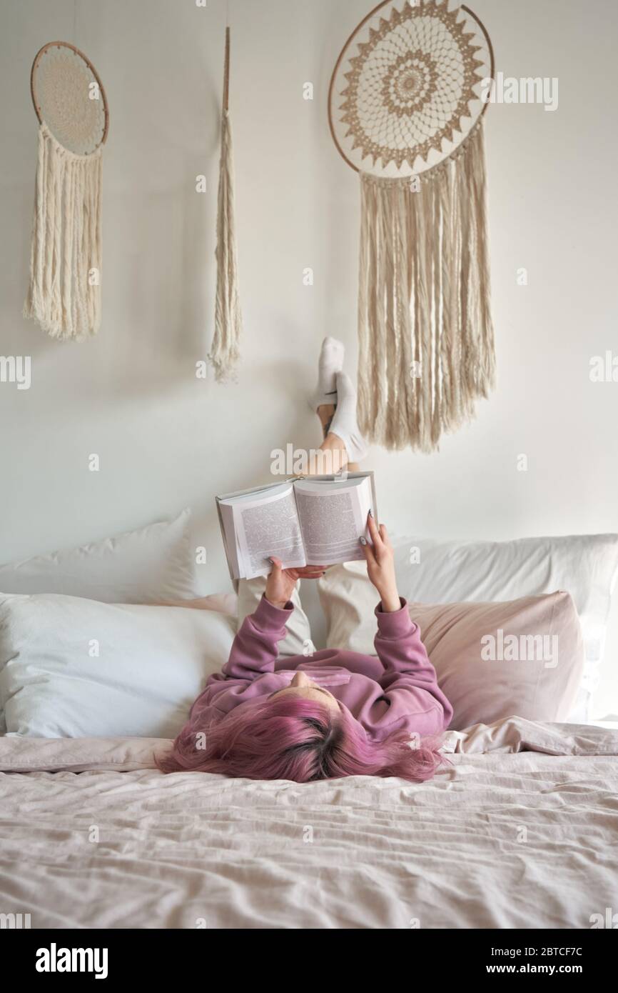 Teen girl with pink hair lying in bed holding feet up reading book. Stock Photo