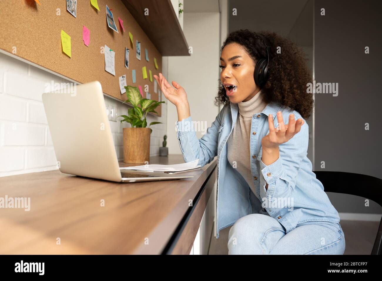 Funny surprised afro american woman wearing headphones video calling on laptop. Stock Photo