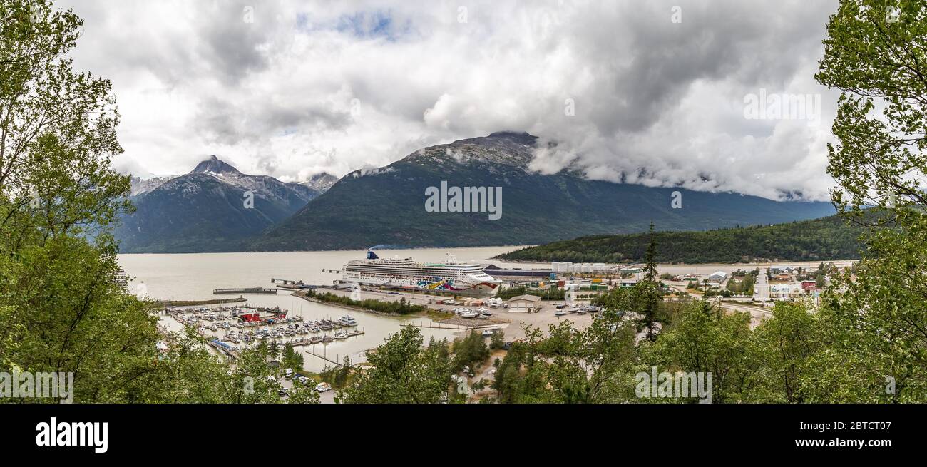 Panoramic view of Skagway port with Norwegian Jewel docked there. Mountains and clouds in the background. Forest in the foreground. Stock Photo