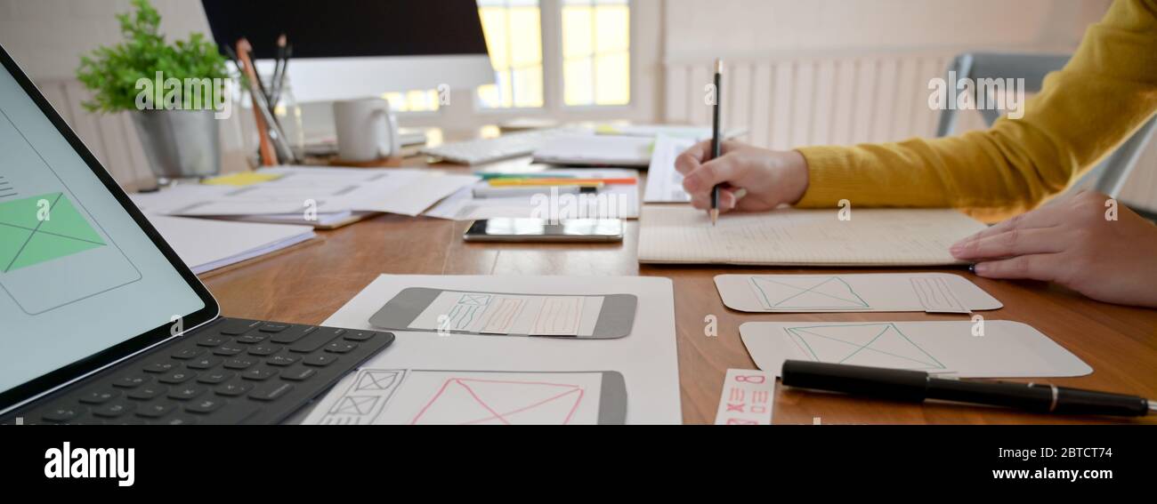 Side view of UI developer working on wooden table with notebook, paperwork and computer Stock Photo