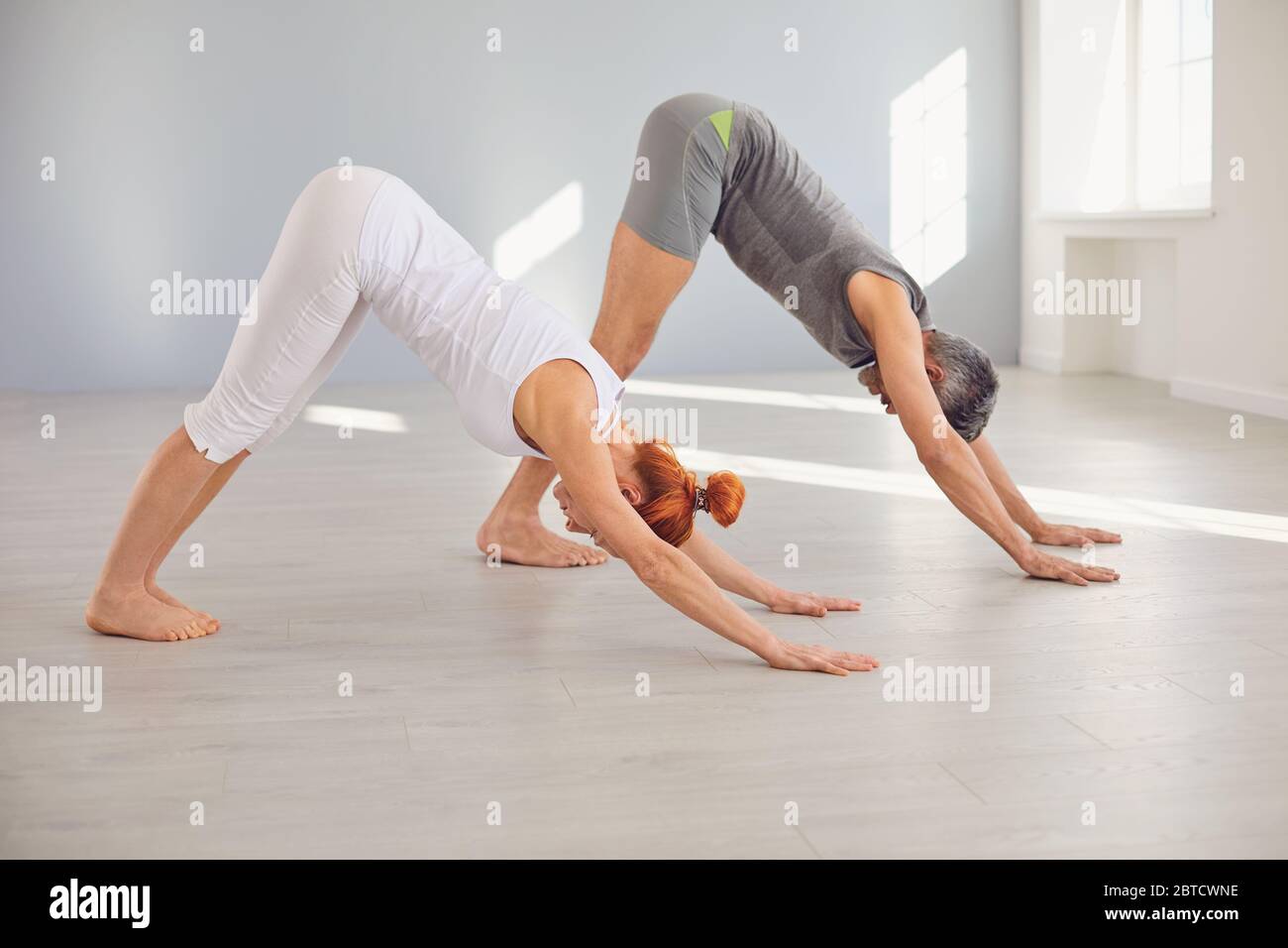 Middle aged man and woman standing in dog position while practicing yoga together in light workout room Stock Photo