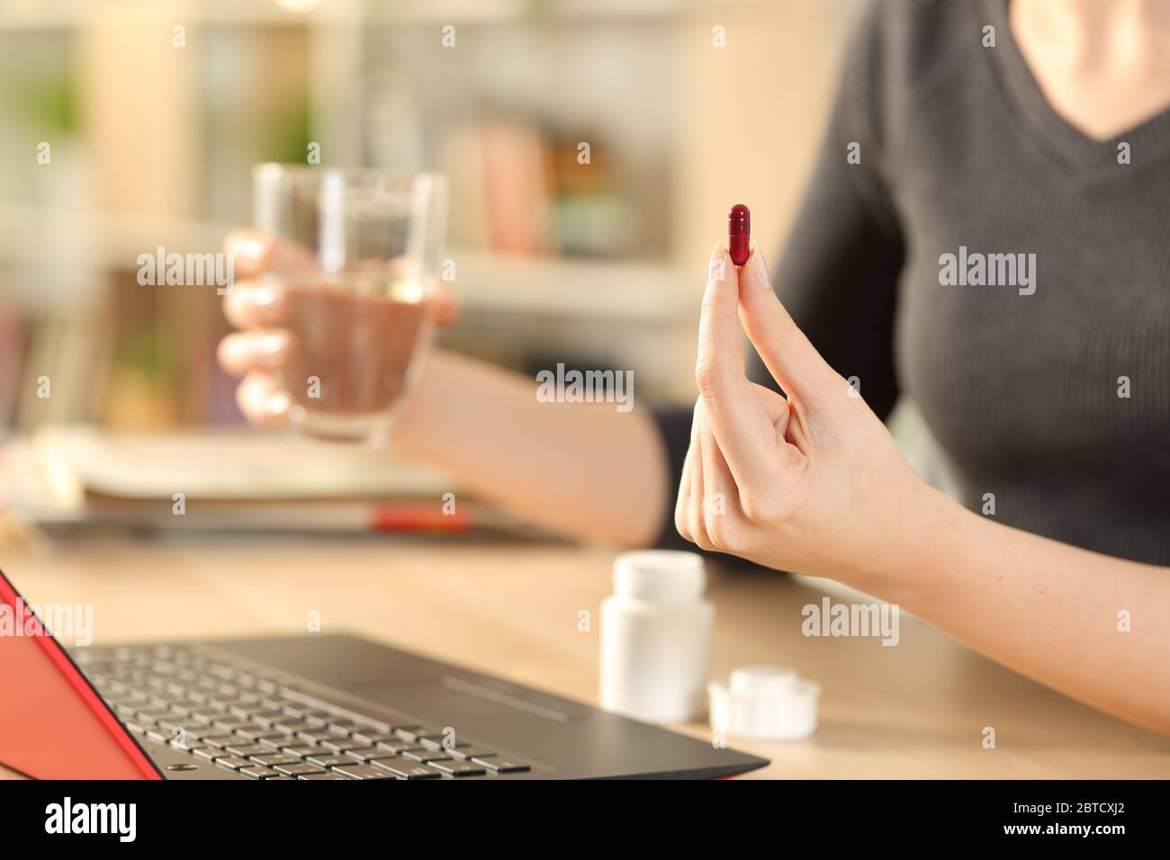Close up of woman hands taking pill holding water glass sitting on a desk at home Stock Photo