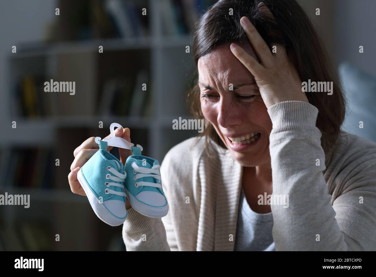 Sad mother crying missing her daughter after miscarriage holding new baby shoes at night at home Stock Photo