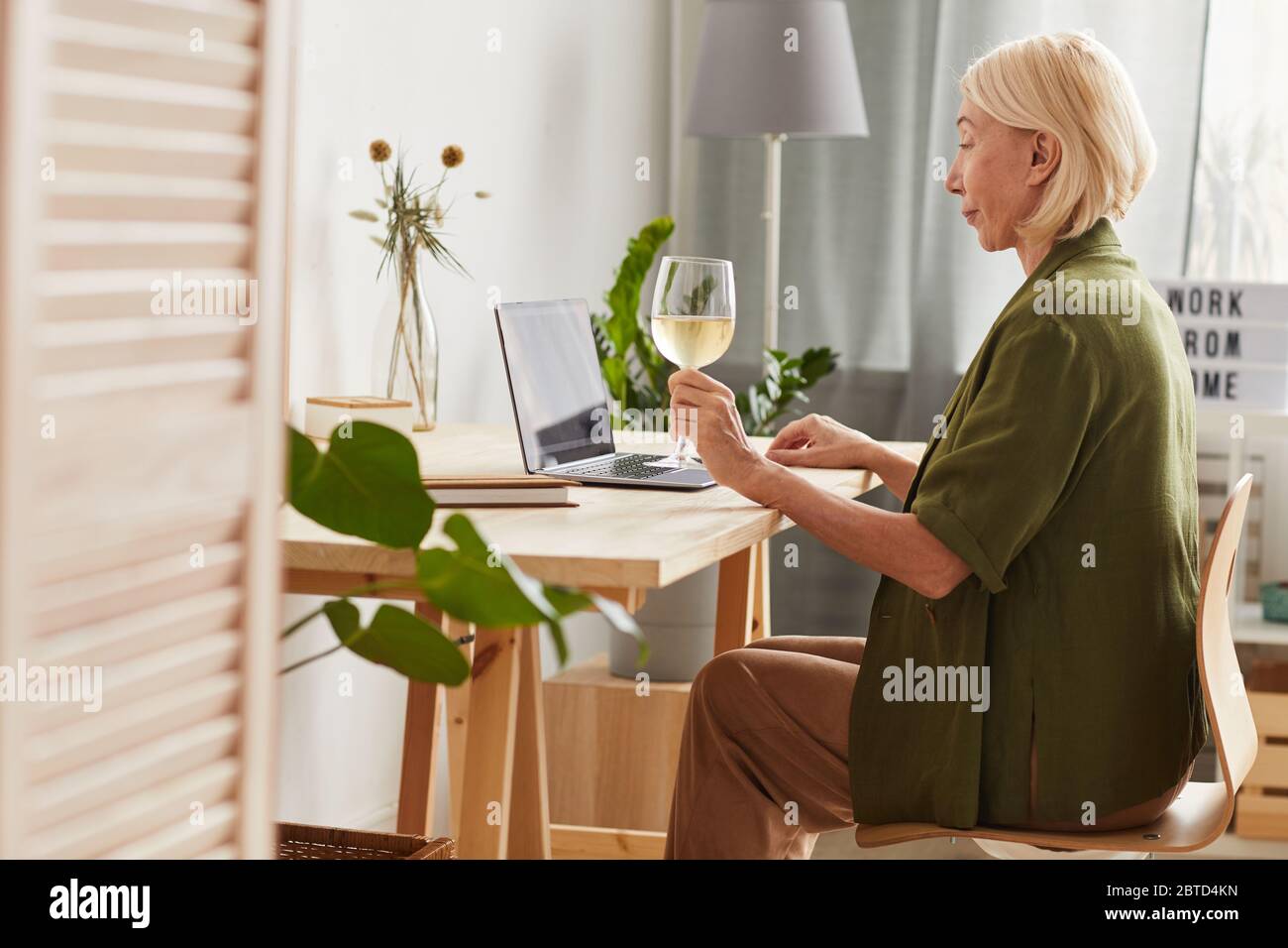 Side view of mature woman sitting at the table with wineglass of wine and using laptop at home Stock Photo