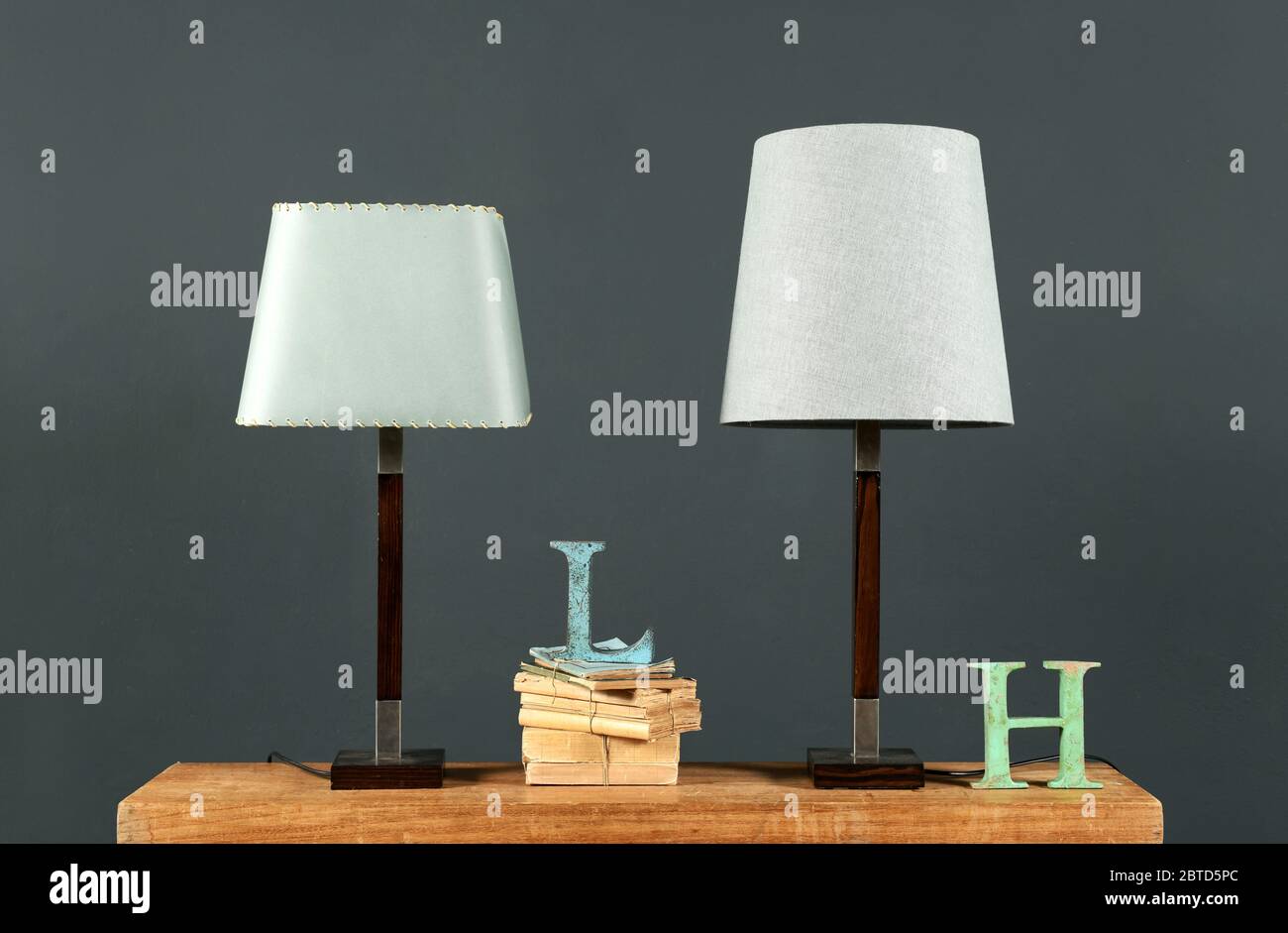 Two vintage table lamps with off white cylindrical shades on an old desk with books and metal letters against a grey wall Stock Photo