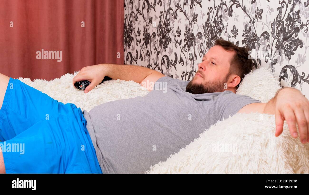 bearded handsome fat man is watching TV using the remote control to switch  channels. The male