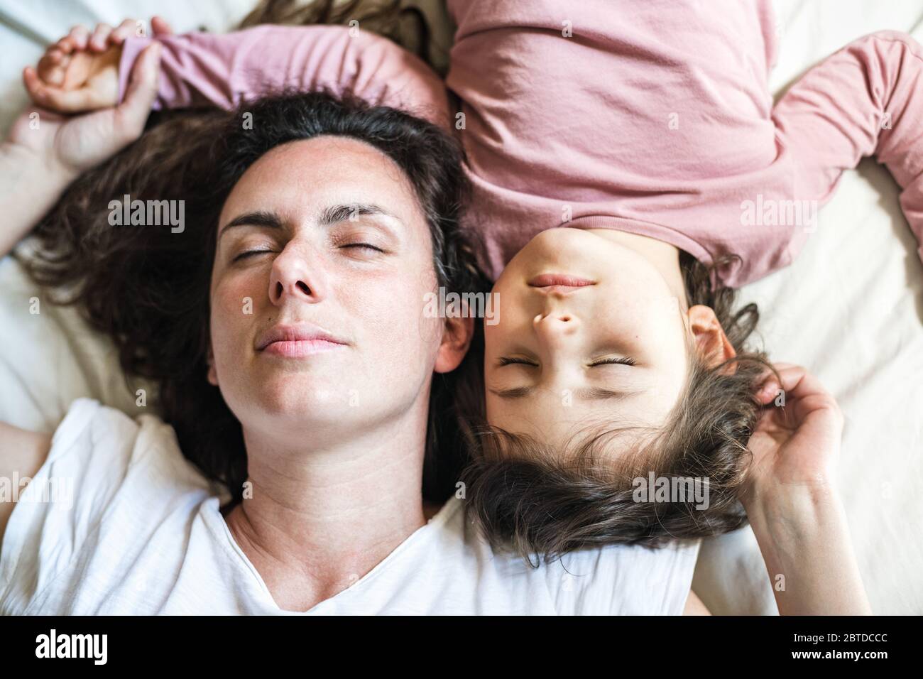 Mother and daughter lie down in bed with their heads facing up. Their eyes are closed having a moment of peace and connection Stock Photo