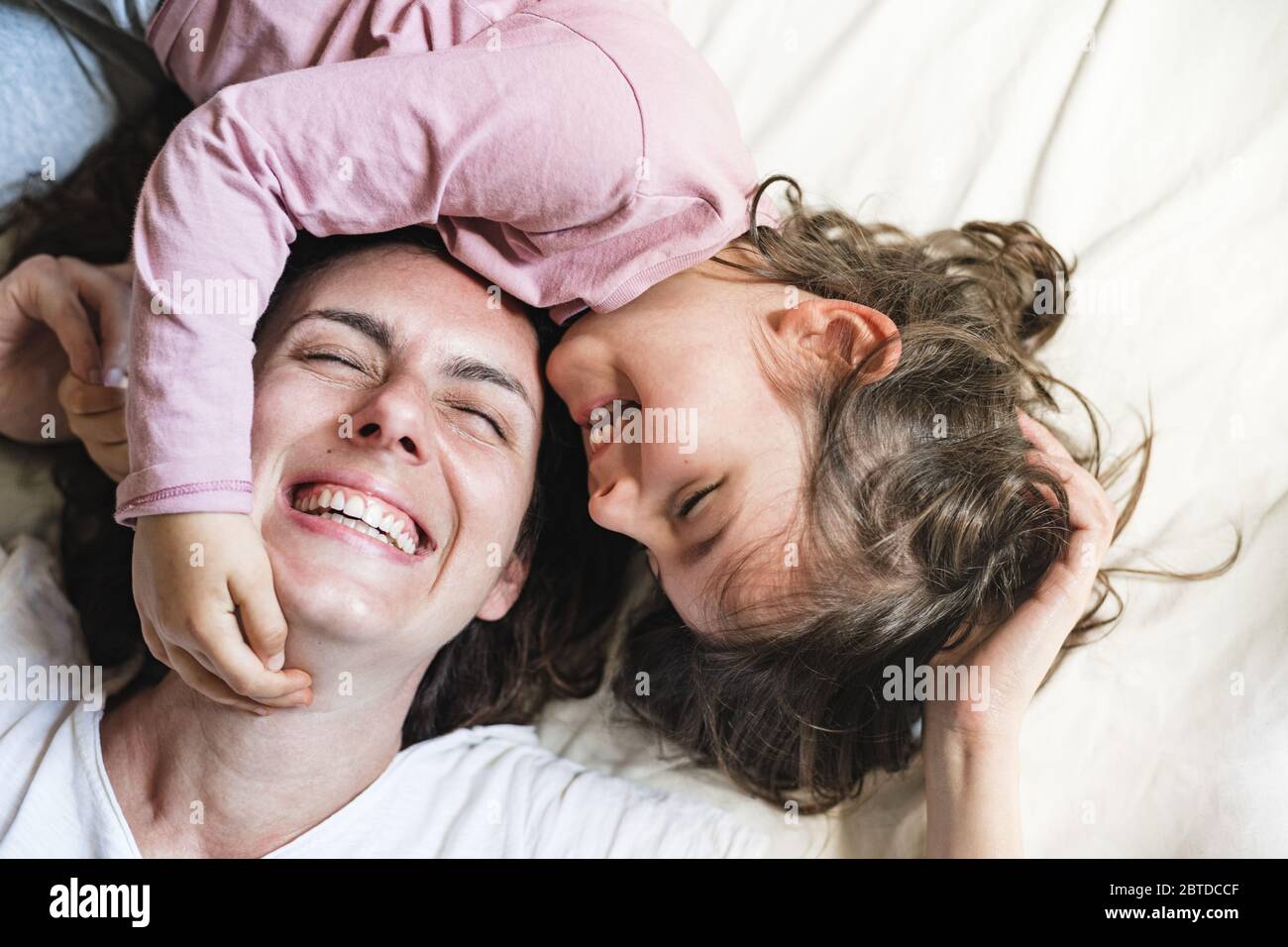 A little girl lays down in her mother's bed as she grabs her face and laughs. They are having a great time playing around Stock Photo