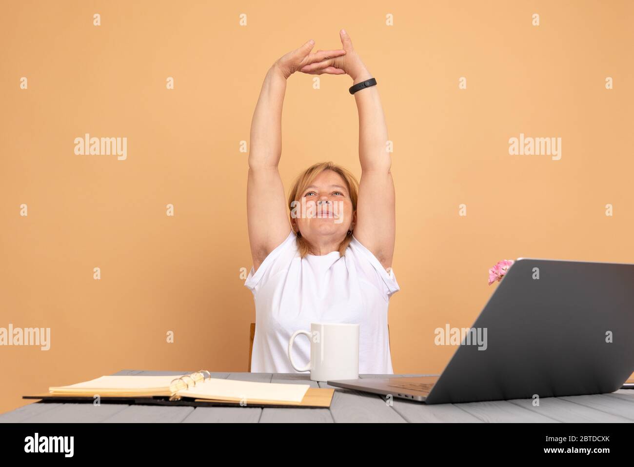 Senior woman sitting at the table with laptop, resting with raised hands, showing hairy unshaven female armpits. Body positive trend Stock Photo