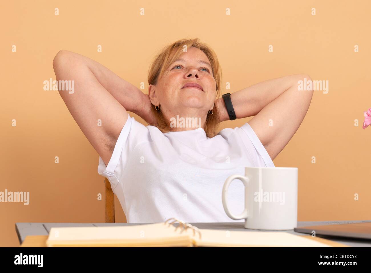 Middle age woman in white t-shirt sitting at the table, holds her hands behinde her head and shows hairy unshaven female armpits Stock Photo