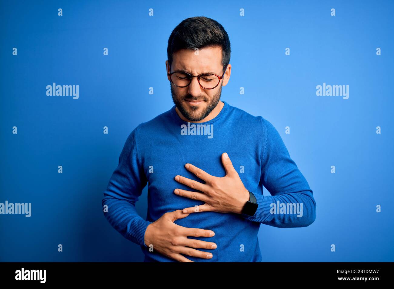 Young handsome man with beard wearing casual sweater and glasses over blue background with hand on stomach because indigestion, painful illness feelin Stock Photo