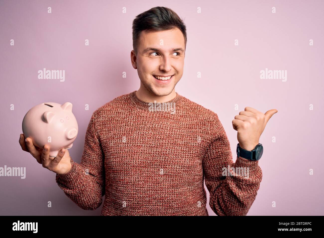 Young handsome caucasian man holding piggy bank for savings over pink ...