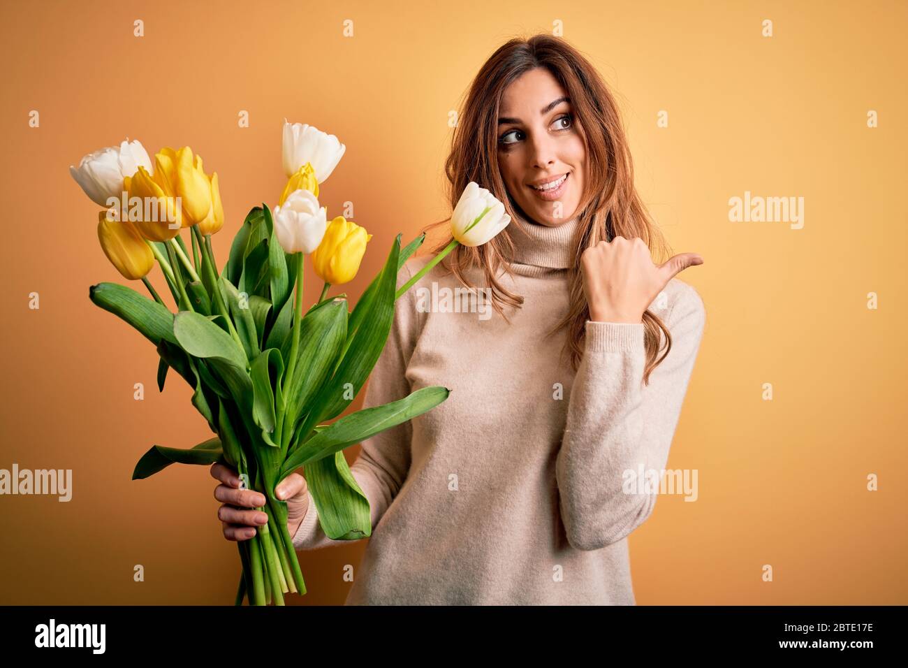 Young beautiful brunette woman holding bouquet of yellow tulips over isolated background smiling with happy face looking and pointing to the side with Stock Photo