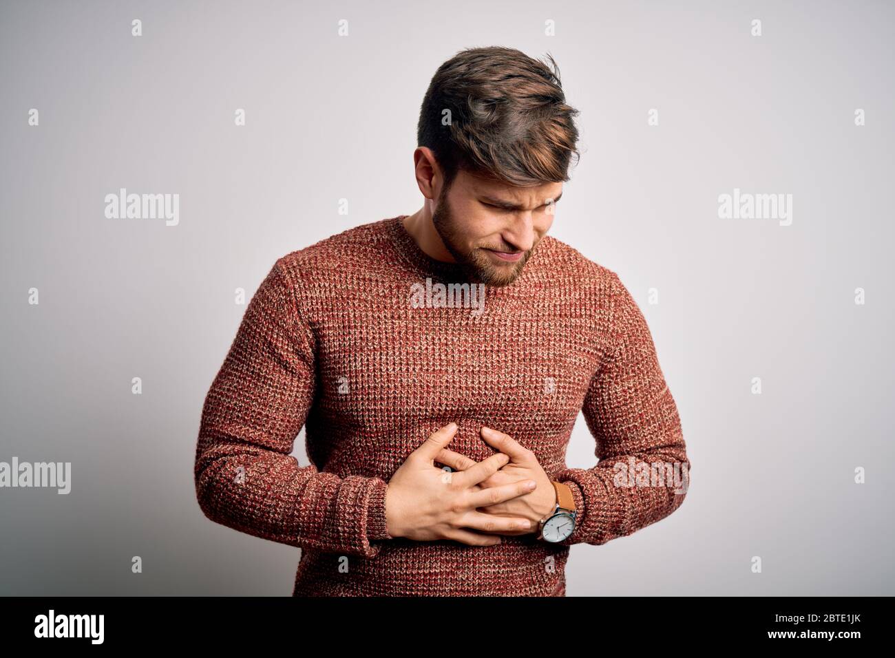 Young blond man with beard and blue eyes wearing casual sweater over white background with hand on stomach because indigestion, painful illness feelin Stock Photo