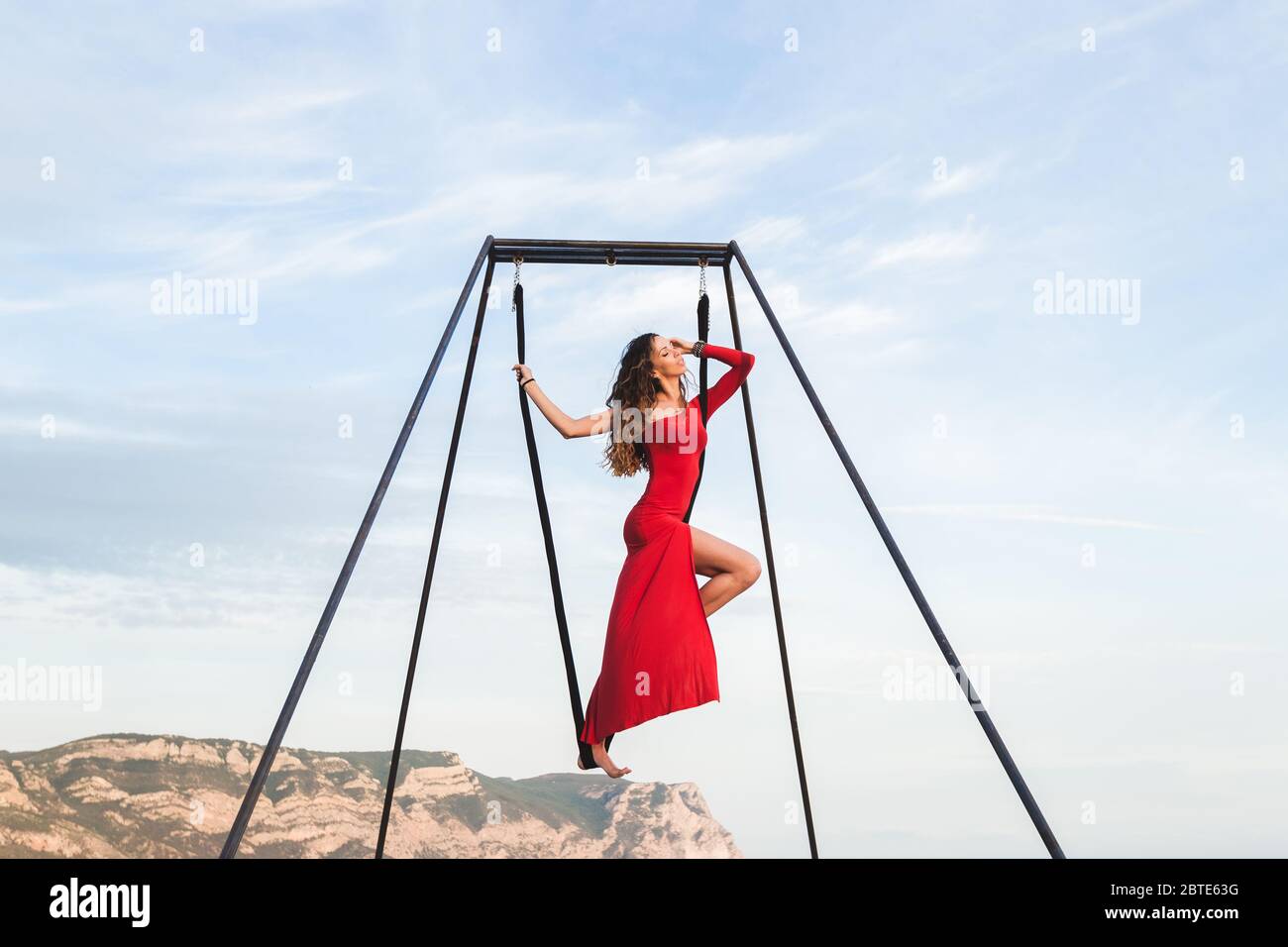 Woman in red dress practicing pole fly dance poses in a hammock outdoor with a mountain view. Female sports, wellbeing concept. Pilates outdoors. Stock Photo