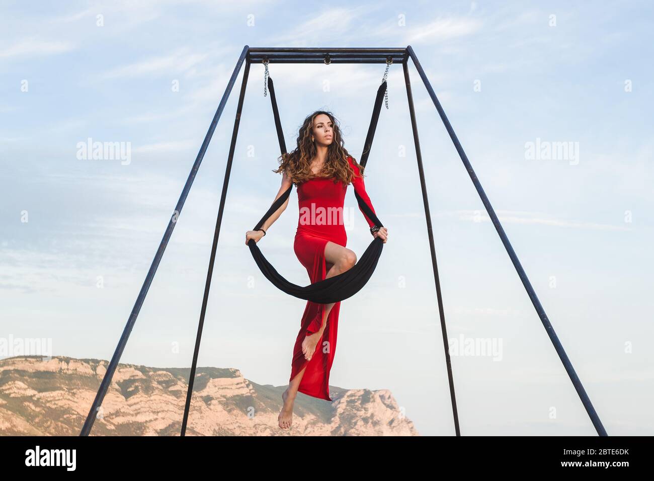 Woman in red dress practicing pole fly dance poses in a hammock outdoor with a mountain view. Female sports, wellbeing concept. Pilates outdoors. Stock Photo
