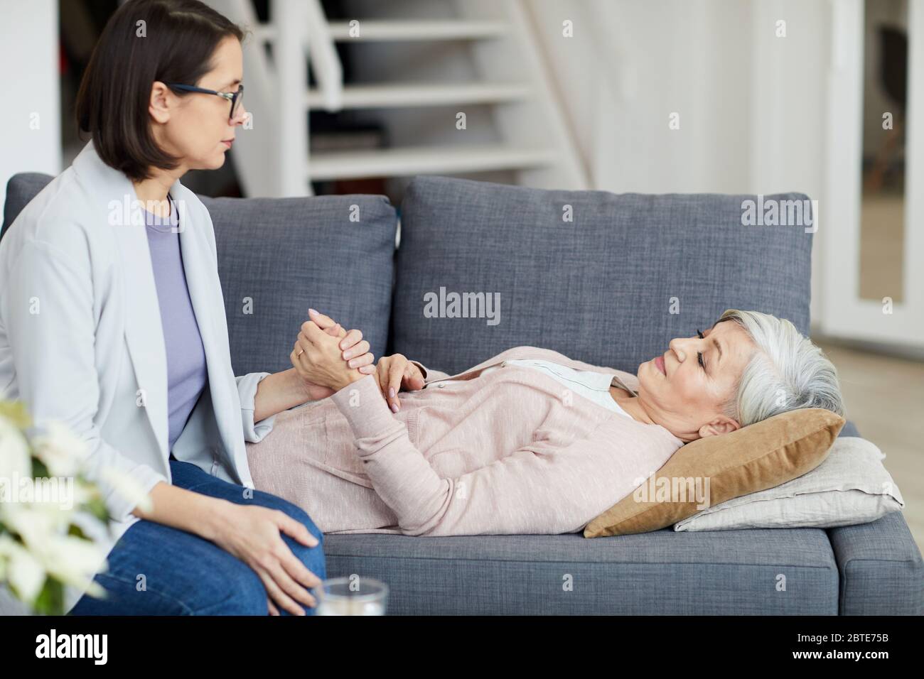 Side view portrait of senior woman lying on couch with young daughter holding her hand and supporting mother, copy space Stock Photo