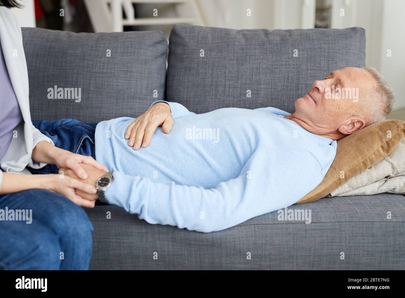 Side view portrait of senior man lying on couch with unrecognizable daughter holding his hand and supporting father, copy space Stock Photo