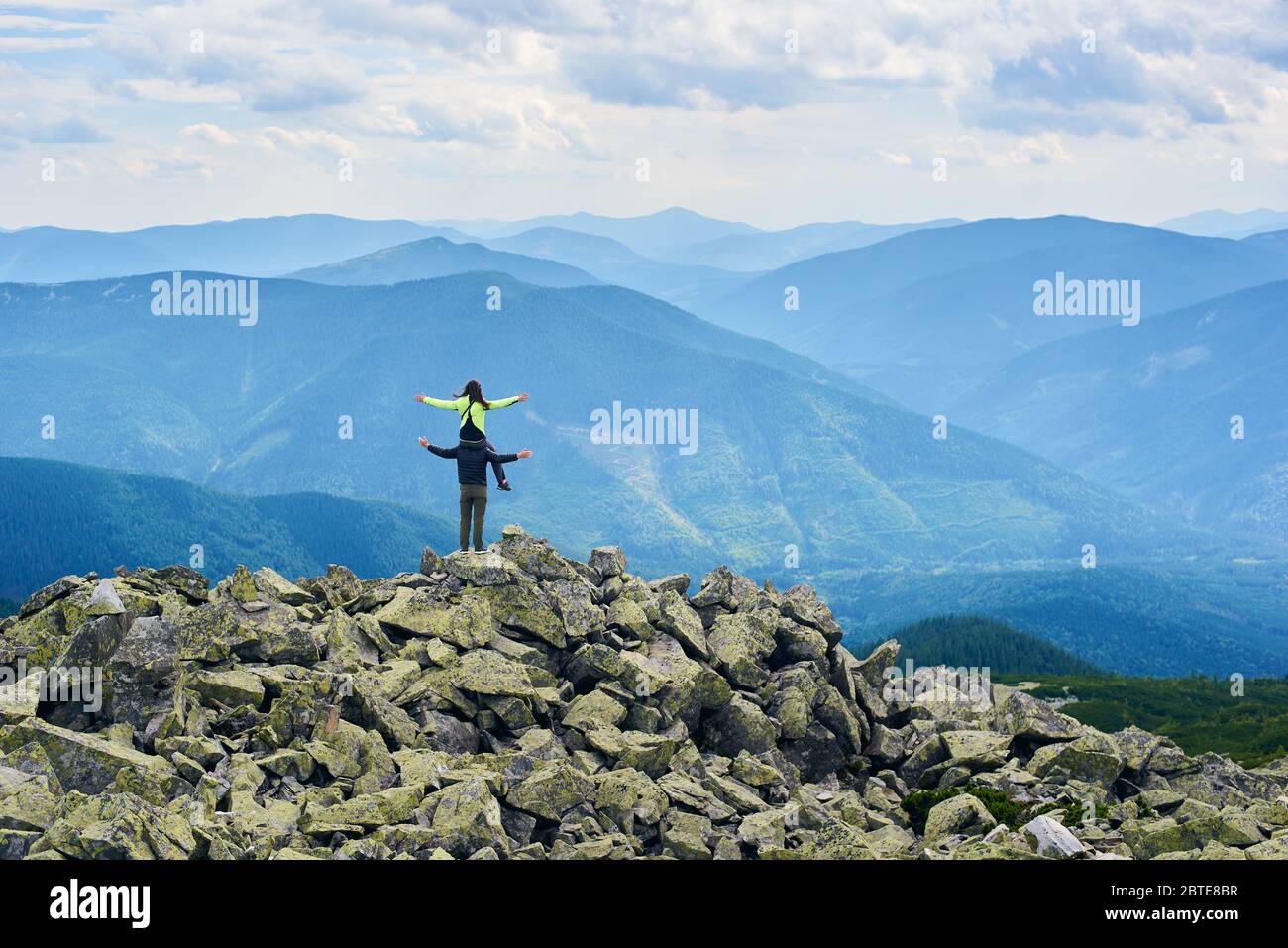 Back view of couple posing, woman sitting on shoulders of man, beautiful mountains scenery on background. Tourists reaching peak together. Wild nature with amazing views. Sport tourism. Stock Photo