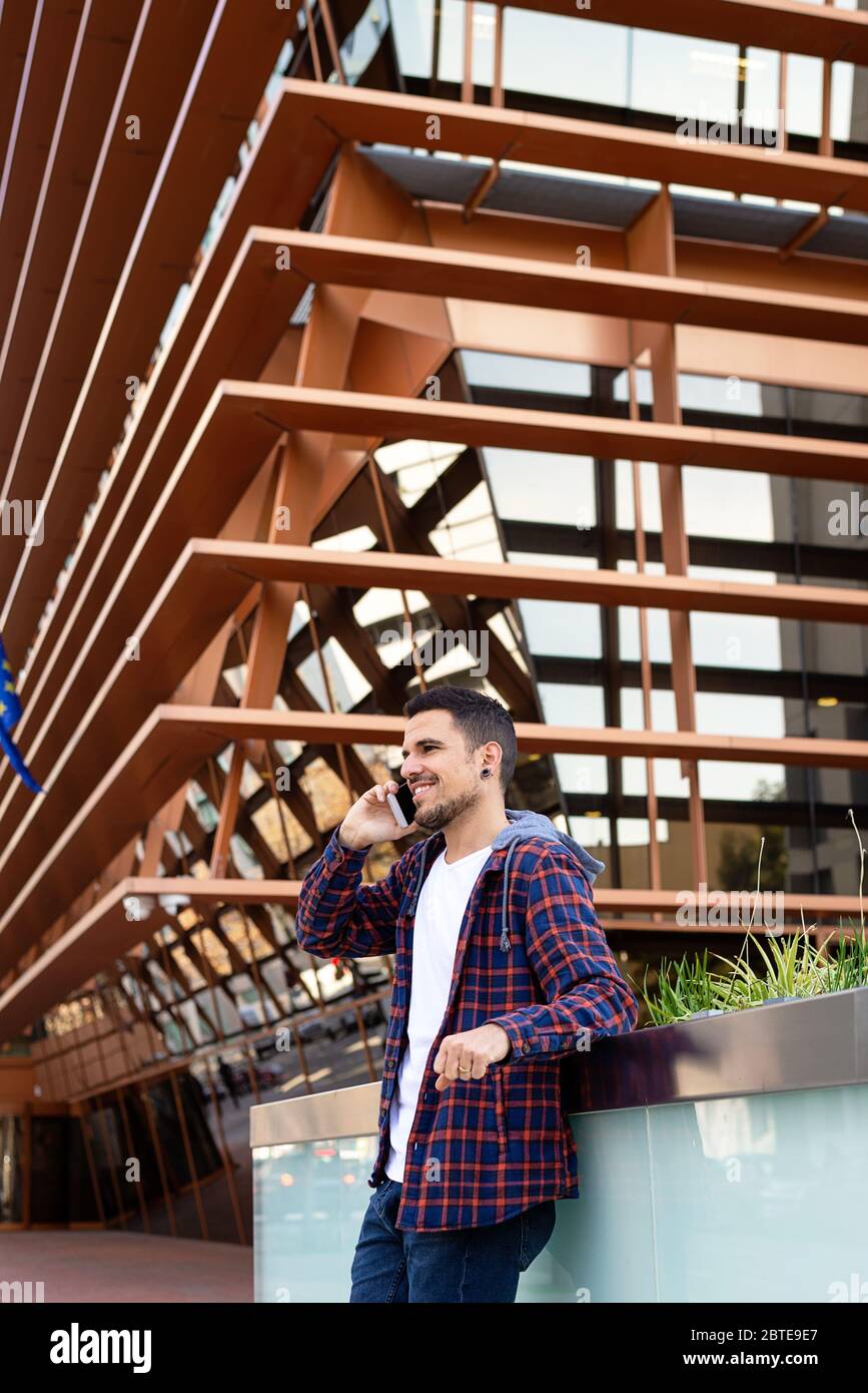 Young bearded male leaning on a wall wearing sunglasses while using a smartphone outside. Stock Photo