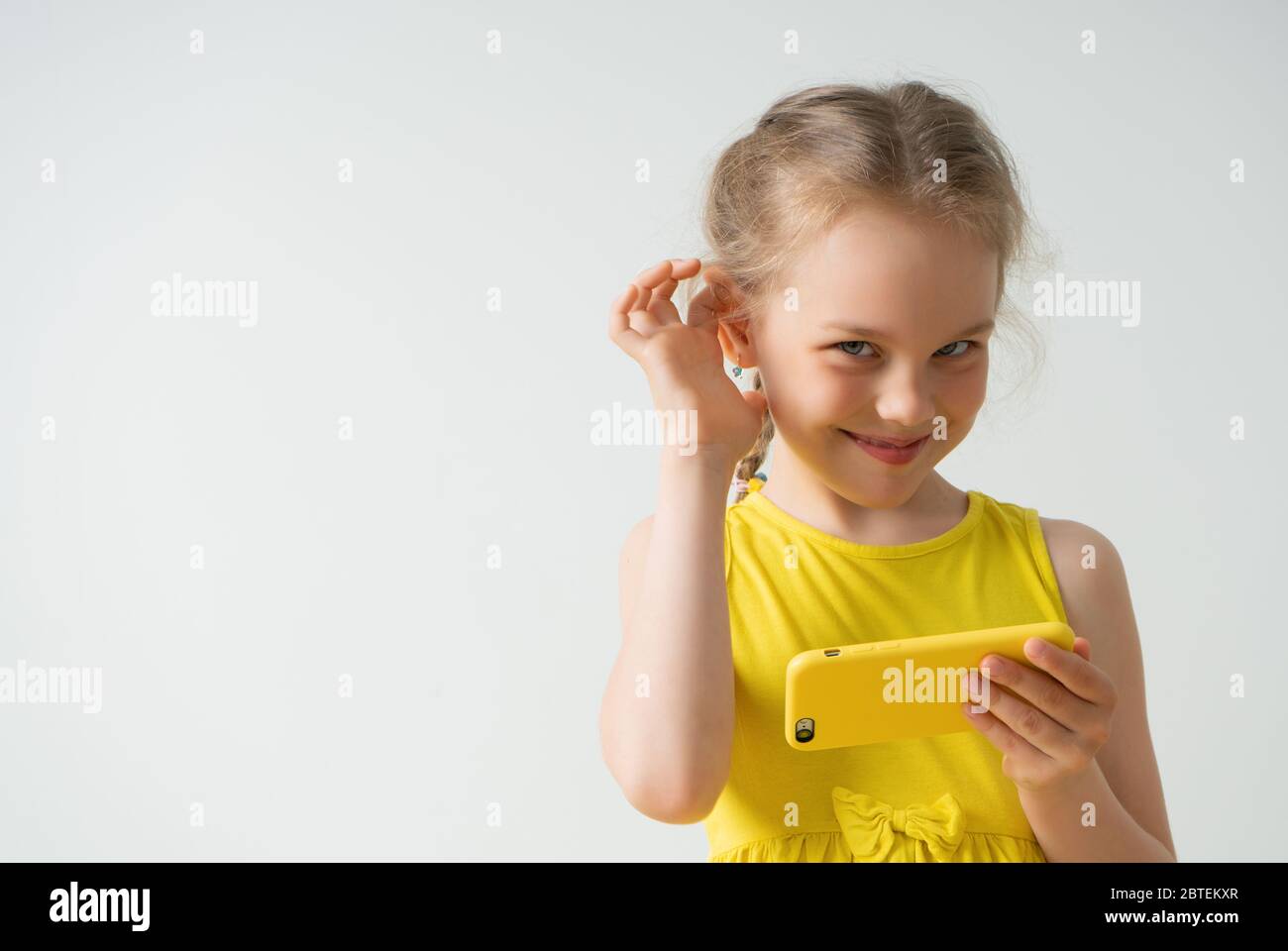 Nice little girl with smartphone looking forward with a sly smile holding hand near ear pretending she does not hear it is enough to play with gadget Stock Photo