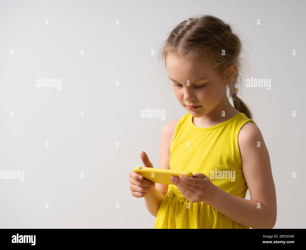 Lovely little girl in bright sleeveless dress with cute pigtail enjoys using her stylish smartpthone. Close up studio portrait isolated on white Stock Photo