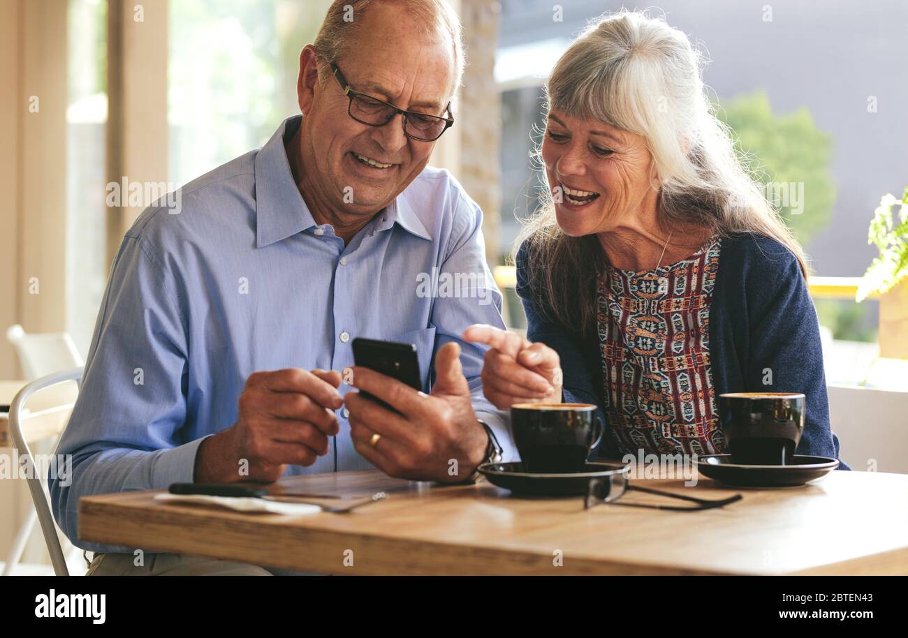 Young senior couple sitting at table in cafe and looking at mobile phone and smiling. Retired man and woman in coffee shop using smart phone. Stock Photo
