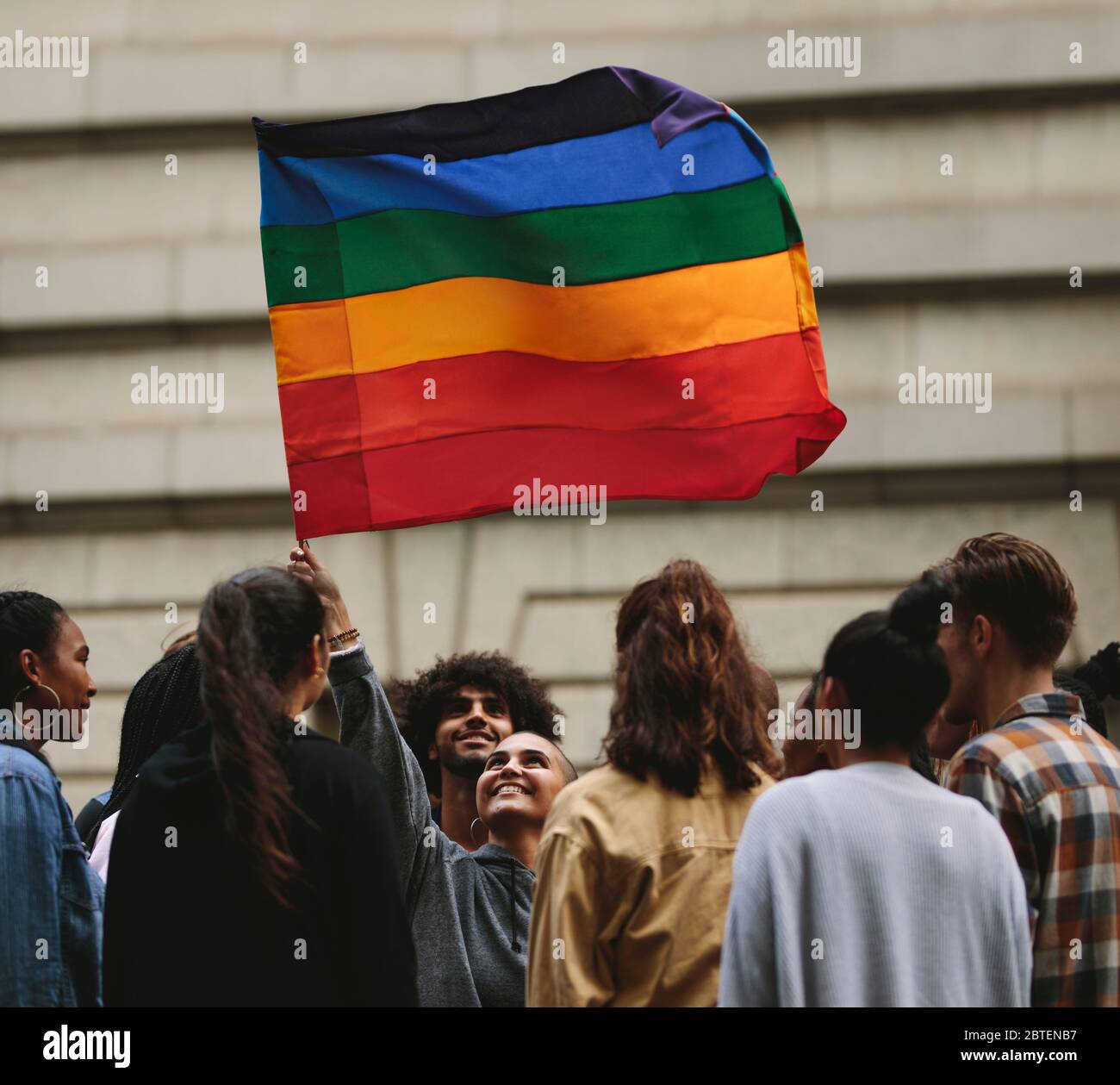 People participate in the annual Pride Parade and celebrations in the city. Young woman waving gay rainbow flag with people standing around. Stock Photo