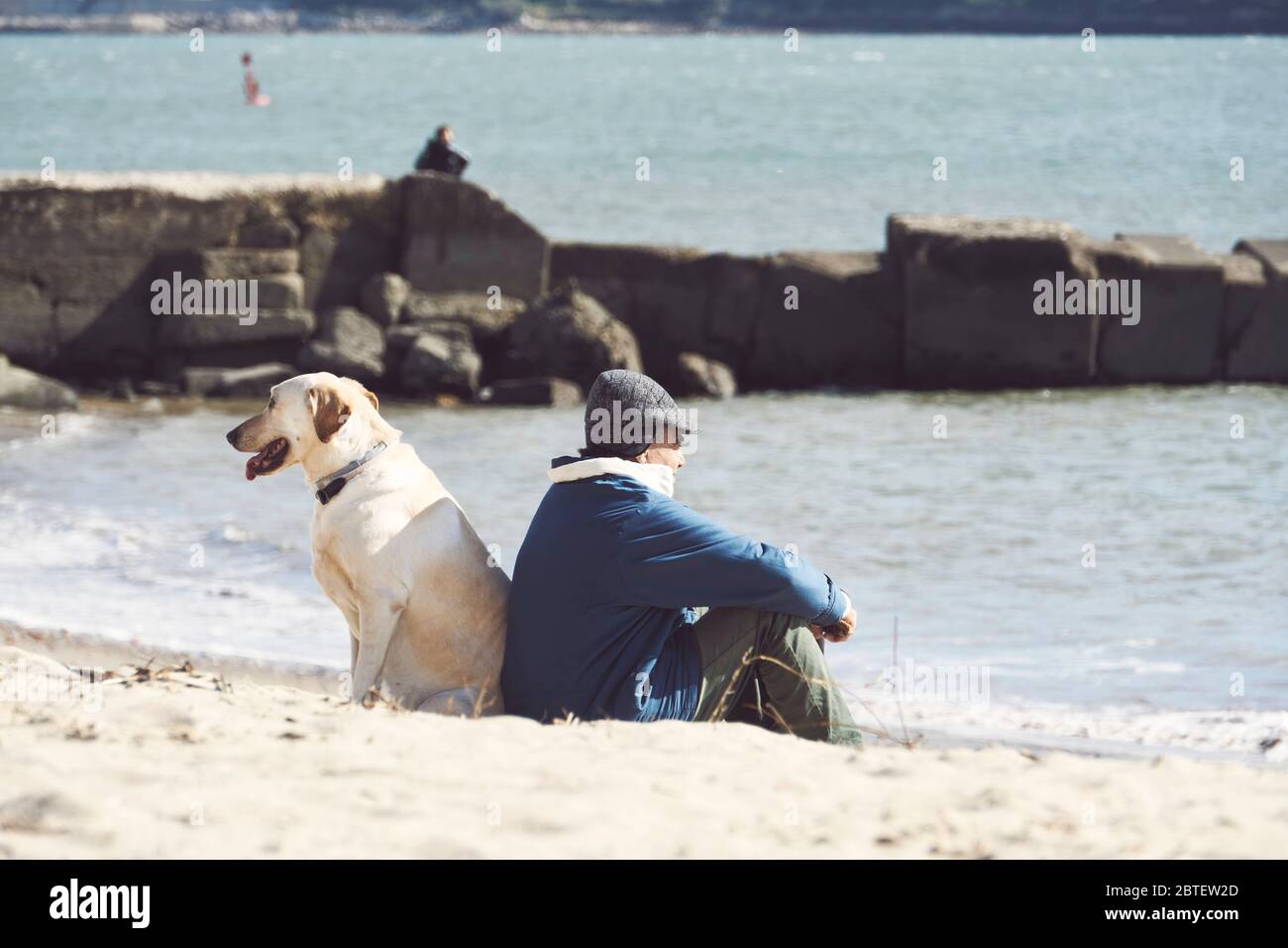 Old man and his dog sitting back to back on a small beach in Lisbon, Portugal, man looking to the sea, looks like a fisherman Stock Photo