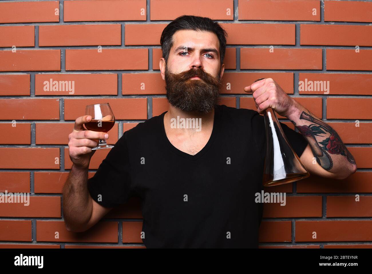 Man with beard and mustache holds alcoholic beverage on brick wall background. Guy with glass and bottle of cognac. Macho with confident face drinks brandy or whiskey. Service and catering concept. Stock Photo