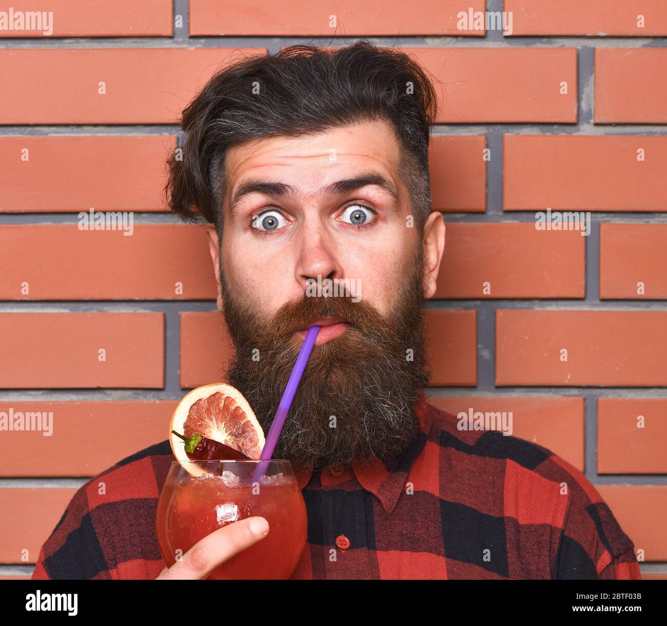 Alcohol and spirit concept. Hipster enjoy strong drink or cocktail. Barman with beard and surprised face drinks out of glass with drinking straw cocktail. Man drinks cocktail on brick wall background. Stock Photo