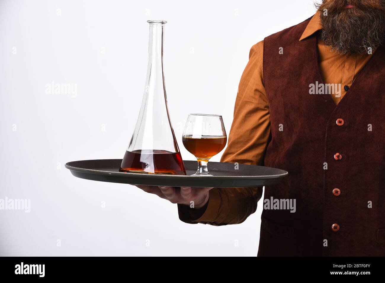 Barman in vintage suede leather waistcoat serves scotch or brandy. Waiter with glass and bottle of whiskey on tray. Restaurant drinks concept. Man with beard holds cognac on white background. Stock Photo