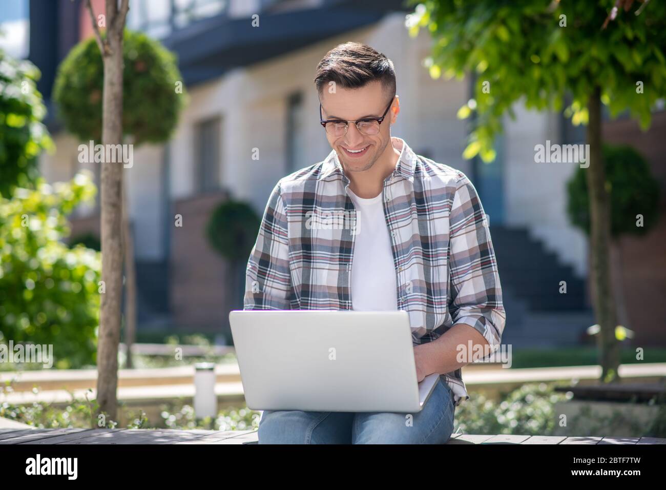 Dark-haired male wearing glasses, sitting in the yard and working on his laptop Stock Photo