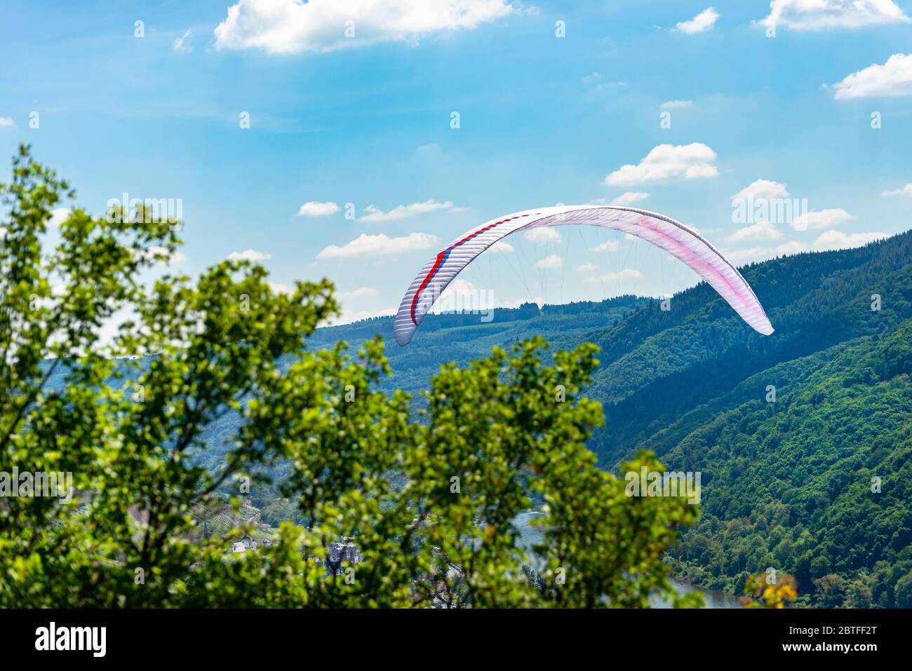 A man flying a white paraglide over the forest and the river on a beautiful sunny day. Stock Photo