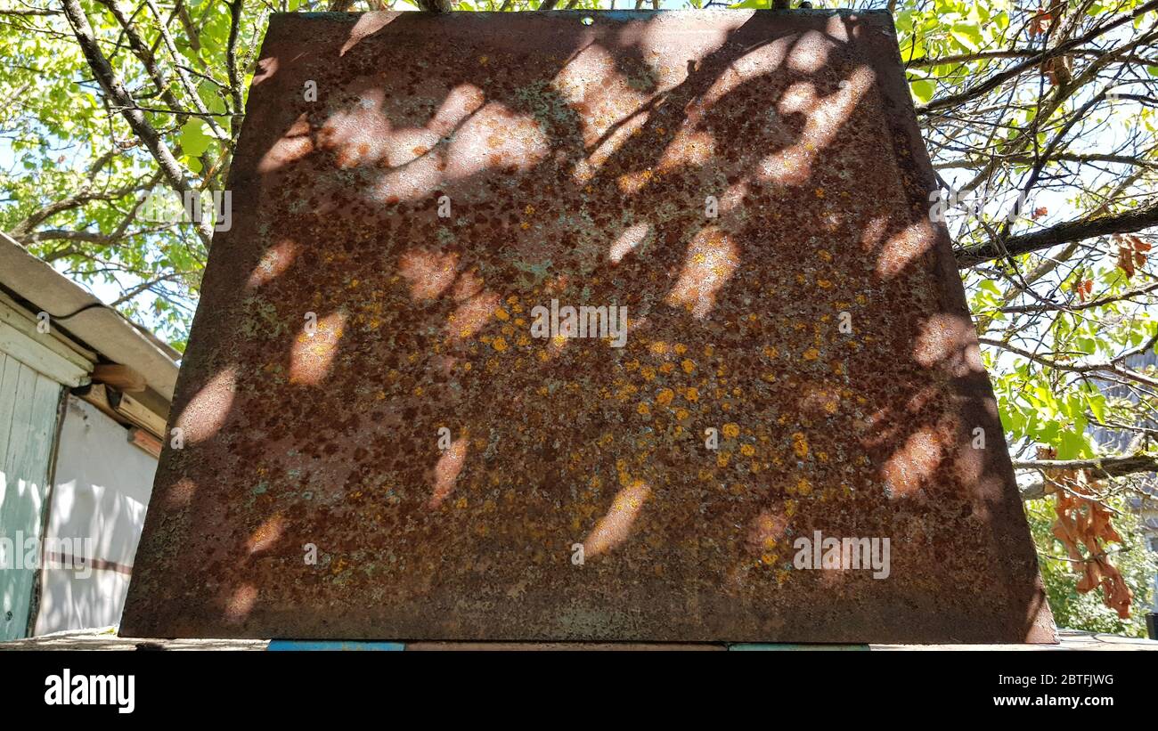 Old weathered brown rusty metal surface with blurry spots of sunlight and shadow. Trapezoid shape sheet of corroded metal with green blurry background Stock Photo