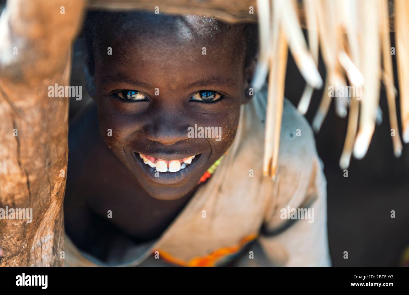 BOYA TRIBE, SOUTH SUDAN - MARCH 10, 2020: Boy optimist with beautiful brown eyes with reflection wearing rags looking out window hole and smiling at Stock Photo