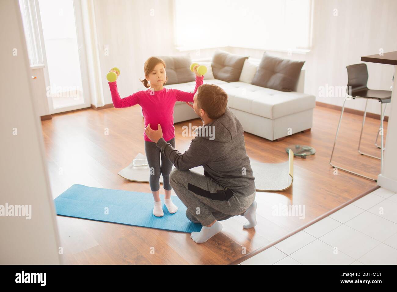 father and daughter are training at home. Workout in the apartment. Sports at home. They are standing on a yoga mat. Girl holding dumbbell. Stock Photo