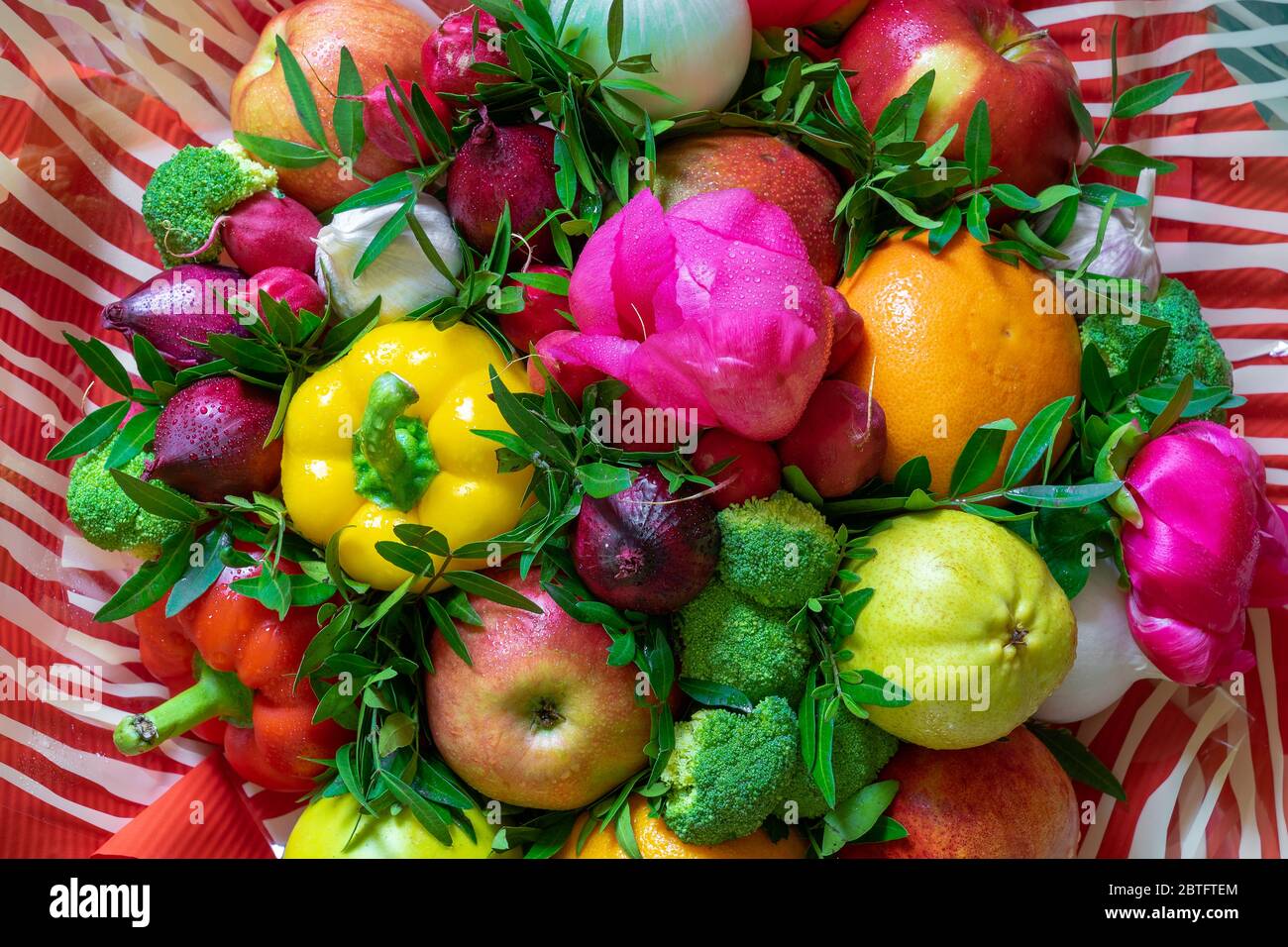 A wonderful multicolored arrangement of a variety fresh fruits and vegetables, pink peonies and green leaves on a red-white striped background Stock Photo