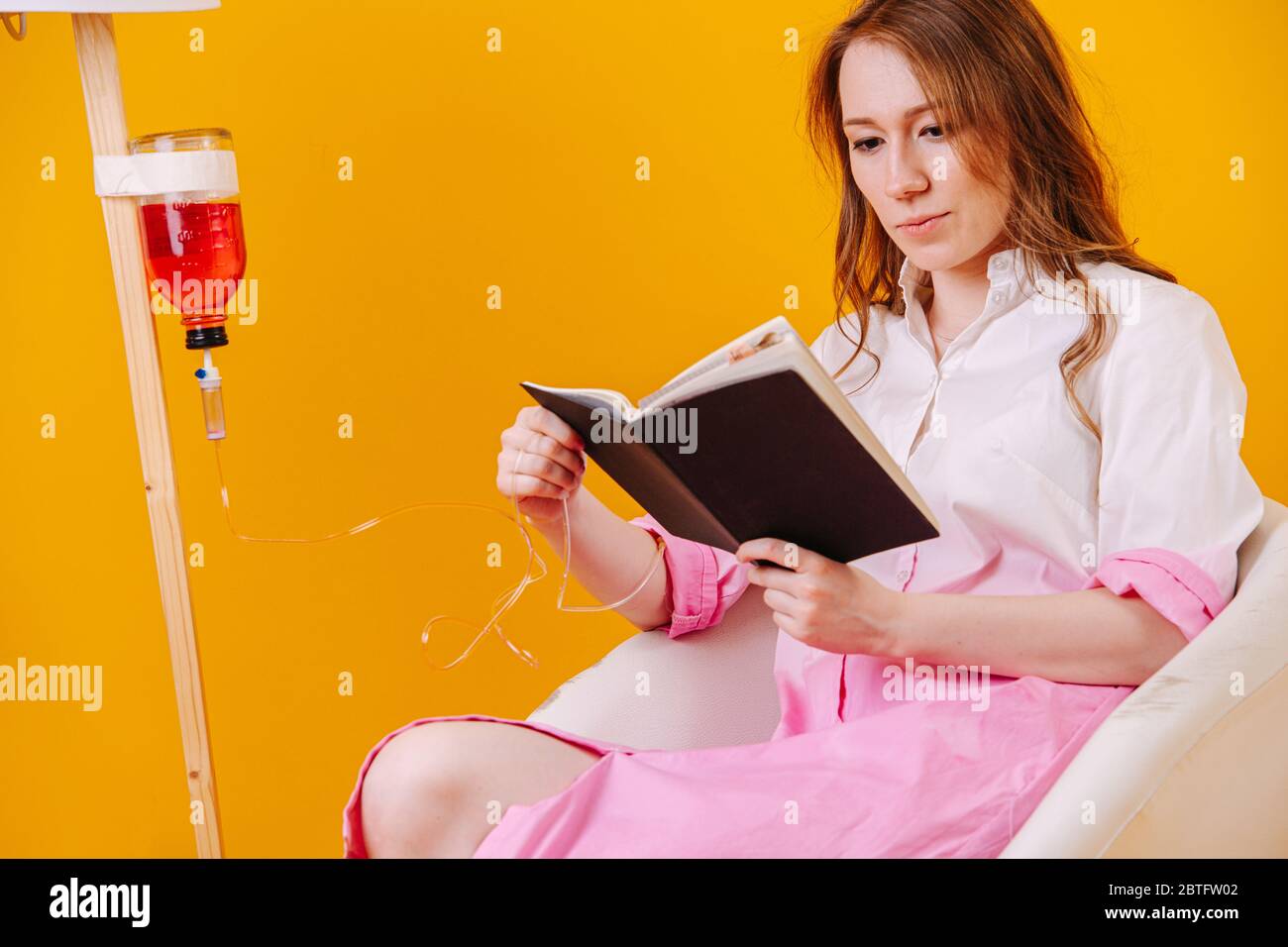 Young sick woman reading while waiting for her infusion drip bottle to go empty Stock Photo