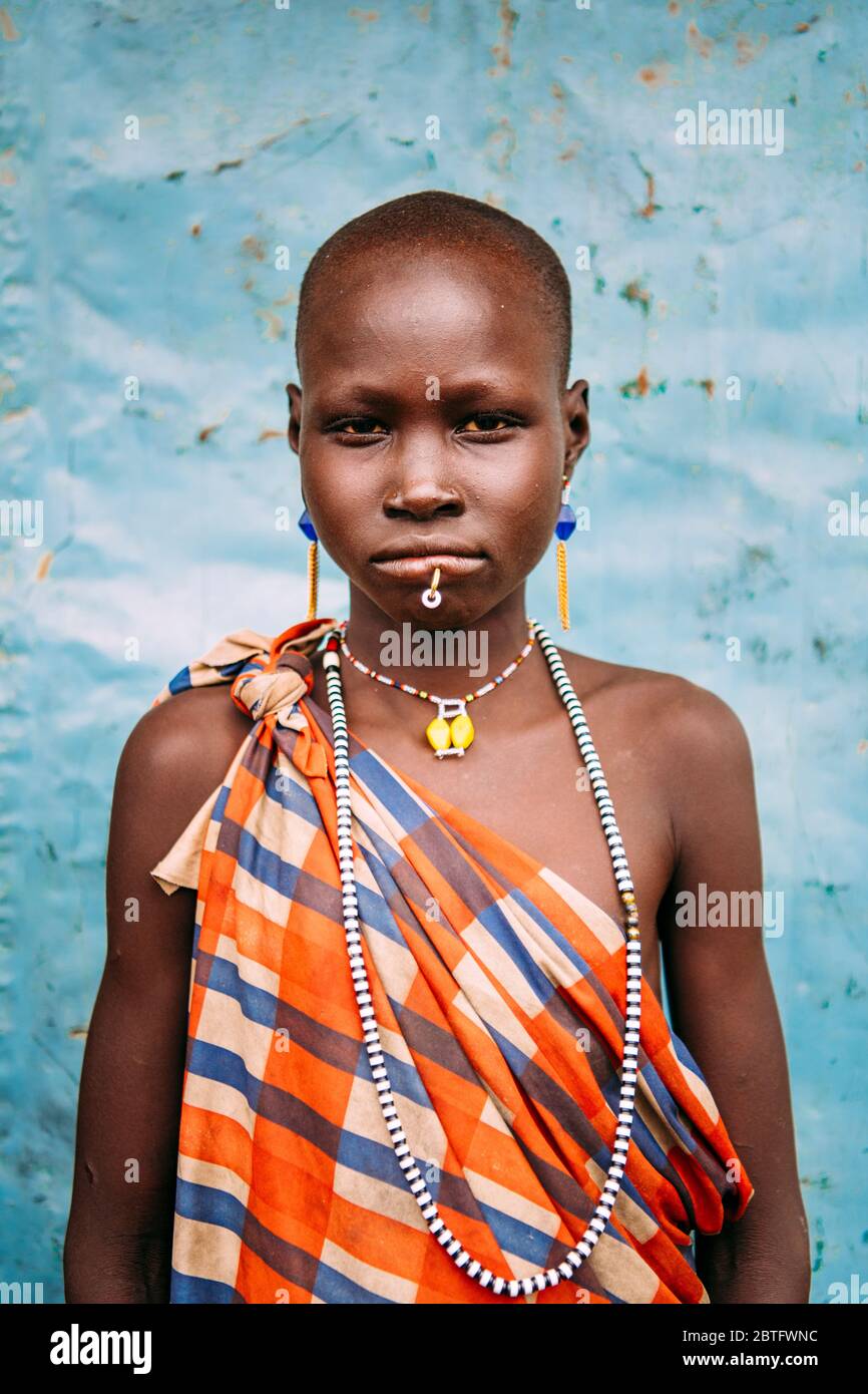 TOPOSA TRIBE, SOUTH SUDAN - MARCH 12, 2020: Girl in checkered colorful garment and with traditional accessories looking at camera against blue wall in Stock Photo