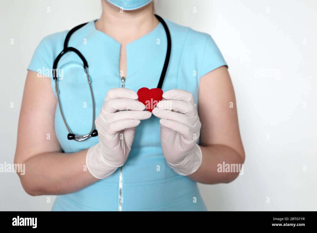 Woman doctor in medical mask holding red knitted heart in hands, saving lives during a covid-19 coronavirus pandemic. Cardiologist, blood donor Stock Photo
