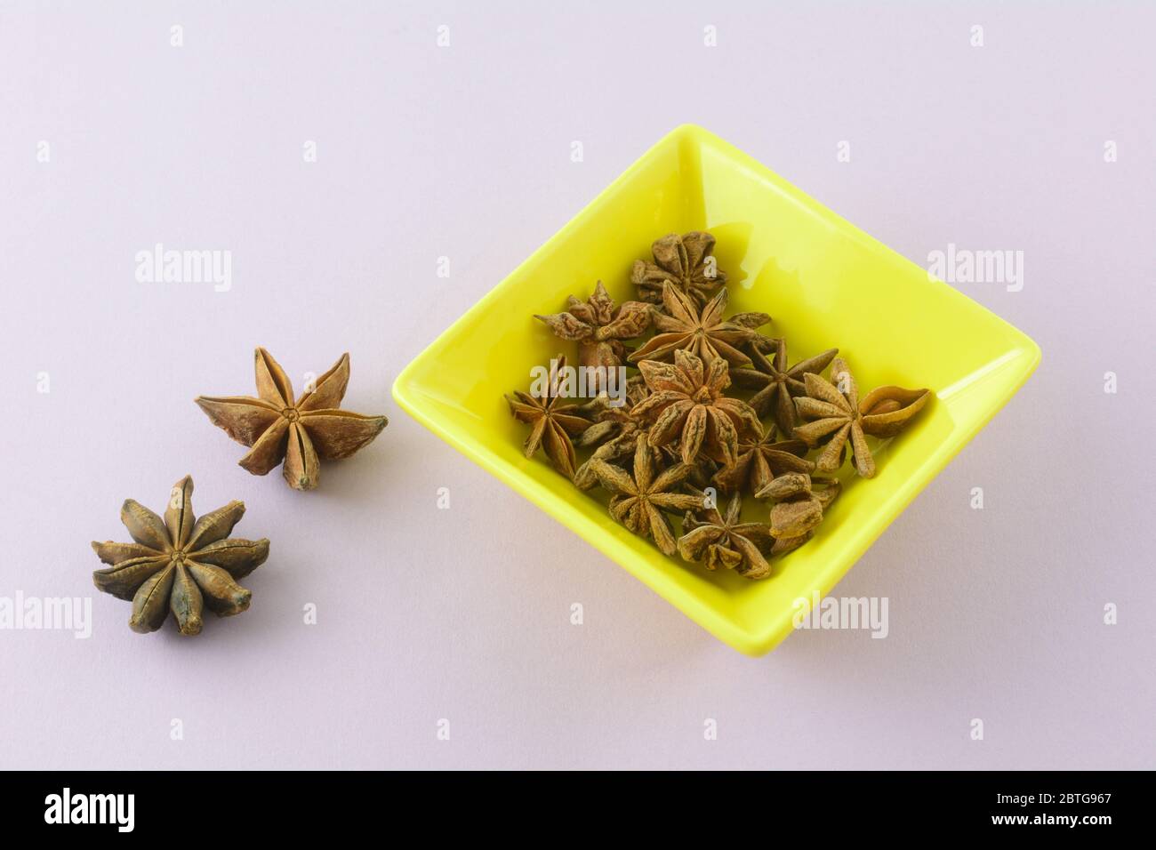 Star anise spice inside and outside small square yellow dish on lavender background Stock Photo