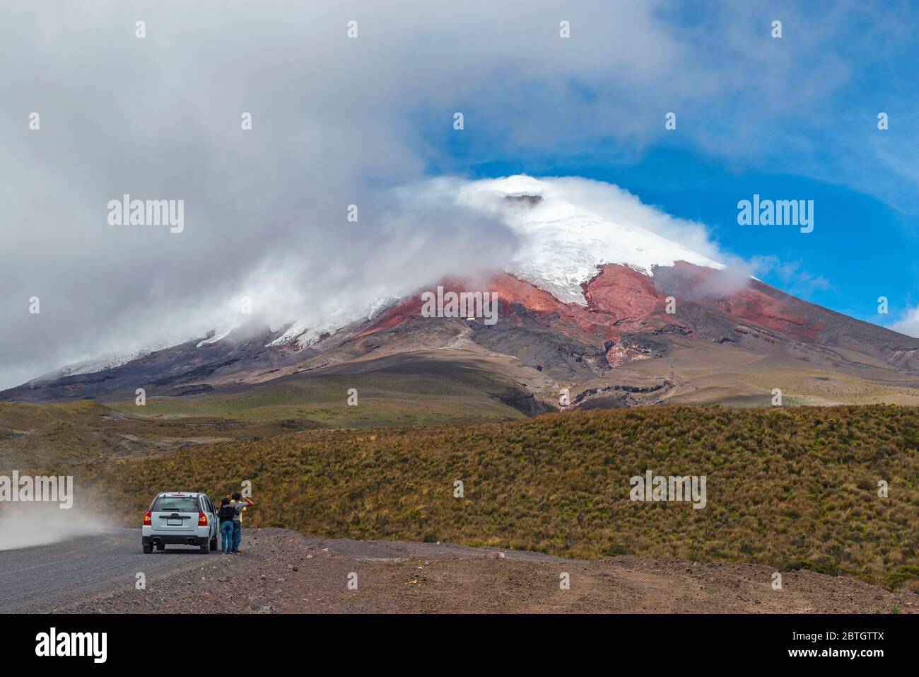 Tourists stopping by a dirt road with their vehicle to enjoy the view over the snowcapped peak of the active Cotopaxi Volcano, Quito, Ecuador. Stock Photo