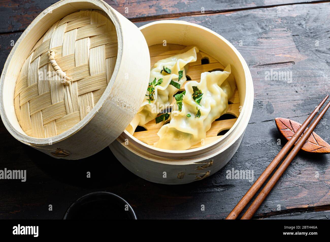 Gyozas potstickers chinese dumplings in wooden steamer with soy sauce fresh herbs and chopsticks on old wood table side view close-up. Stock Photo