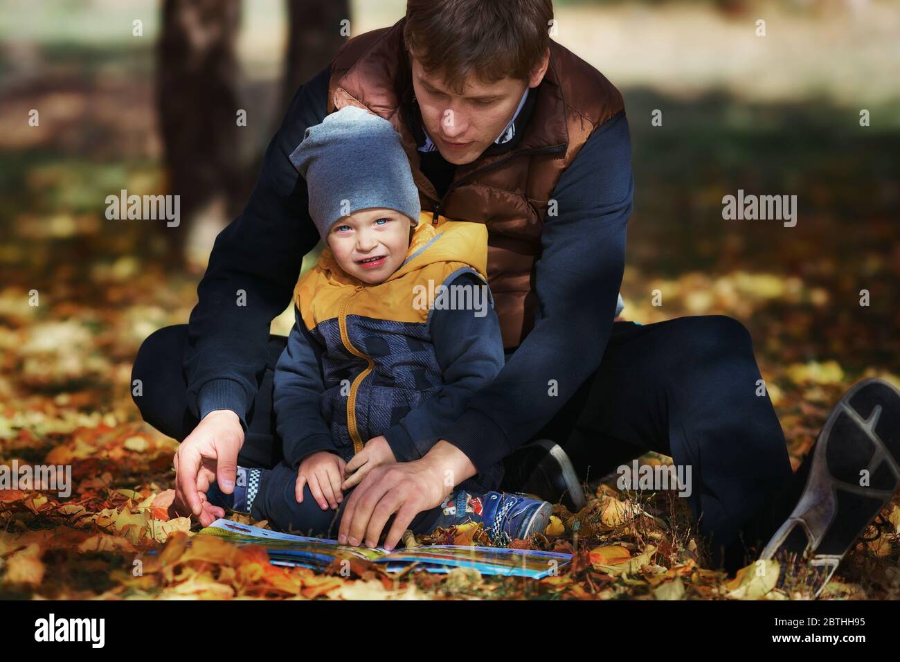 Father's Day. A young father and son in the park on the grass with yellow leaves read a book. Stock Photo