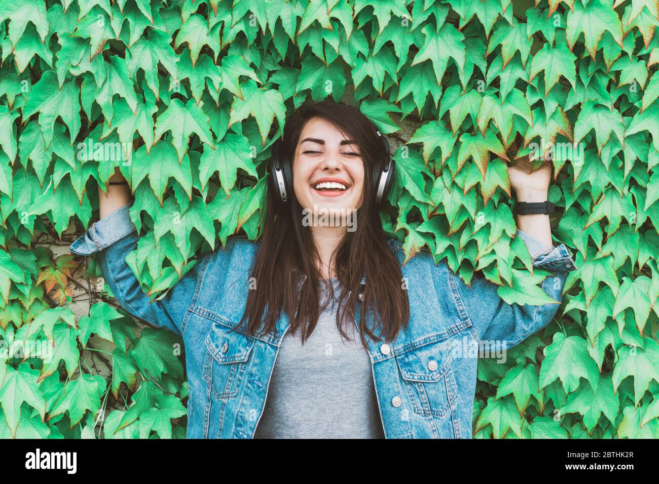 Young hipster girl listens to music leaning against an ivy wall - Pretty woman relaxes with headphones in a city park Stock Photo