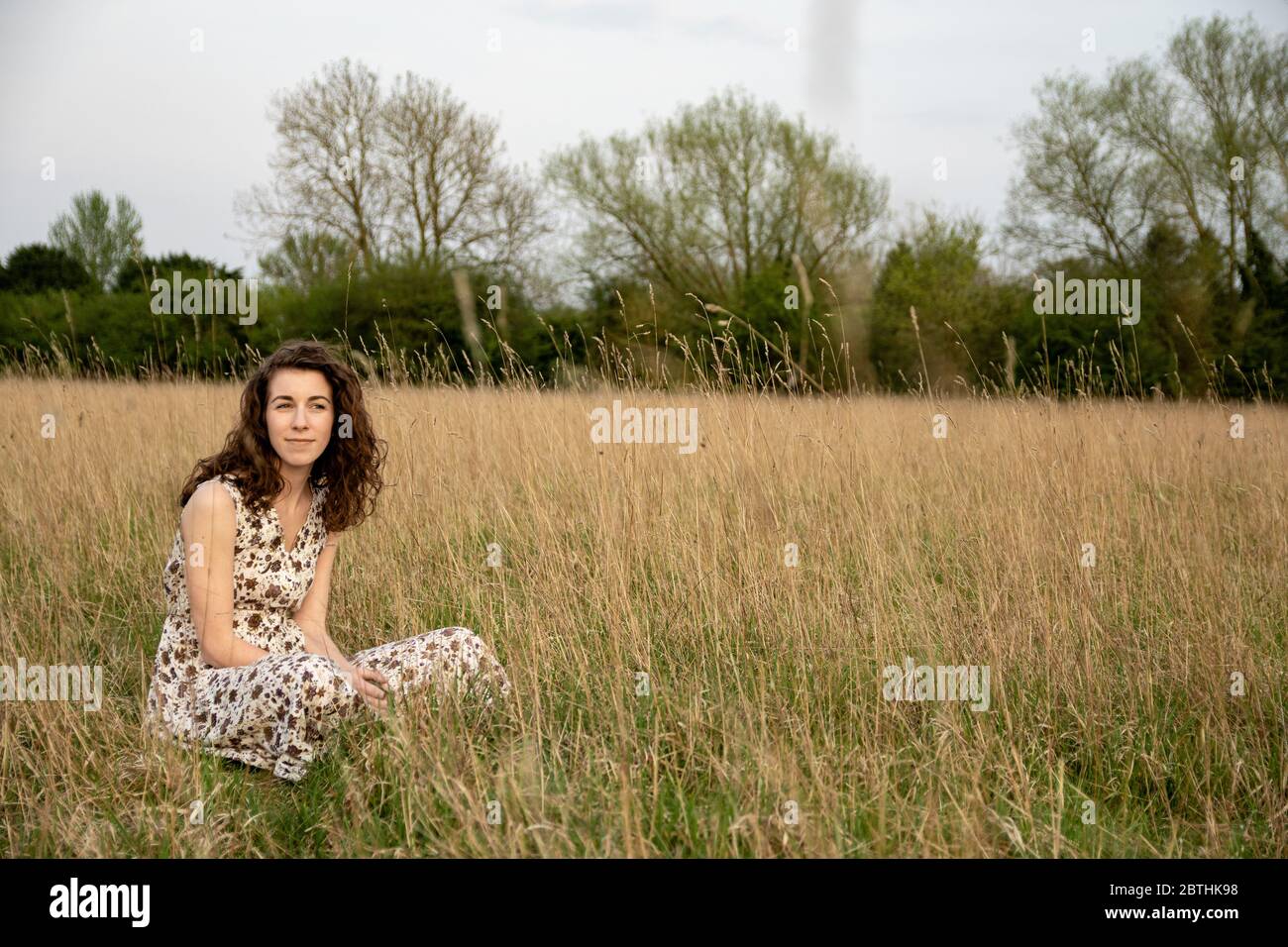 A beautiful and healthy young woman in a dress dances stands peacefully meditating and thinking in a brown grass field at sunset Stock Photo